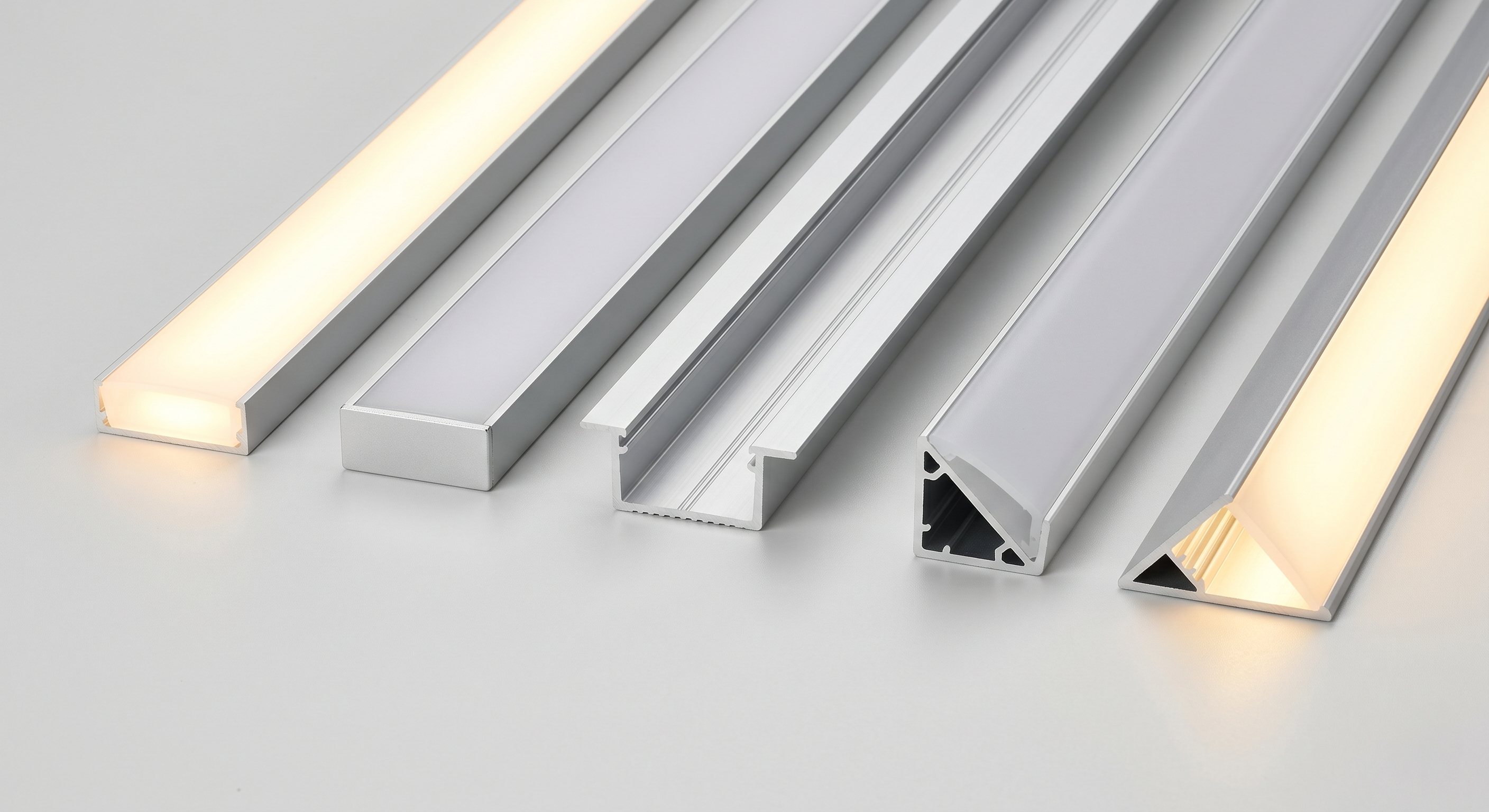 Verschiedene LED-Aluprofile – Aufbau-, Einbau- und Eckprofil – nebeneinander, teils mit warmweiß leuchtendem Diffusor